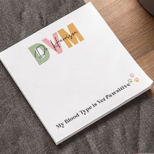 May include: A white 4x4 inch notepad with the text "DVM Dr. Harrison" in green, pink, and orange. The notepad also has the text "My Blood Type is Vet Pawsitive" and paw prints.