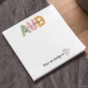 May include: A white 4 x 4 inch notepad with the text "AUD Dr. Harrison" in various colors. The notepad also has the text "Ear to help" and an ear graphic. The notepad is on a dark gray surface.