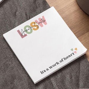 May include: A white 4x4 inch notepad with the text "LOOW Amanda Smith" in pink, green, and orange. The phrase "It's a work of heart" is printed below, with small heart graphics. The notepad is on a dark gray surface.