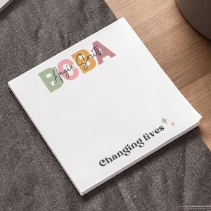 Peut inclure: Un bloc-notes blanc de 10 x 10 cm avec le texte "BOBA Angie Smith" en lettres colorées. En dessous, les mots "Changing lives" sont imprimés en noir, dans une police stylisée. Le bloc-notes est sur une surface grise foncée.