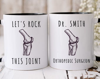 Könnte beinhalten: Zwei weiße Keramiktassen mit schwarzen Henkeln und Rändern. Die linke Tasse zeigt eine schwarze Strichzeichnung eines menschlichen Kniegelenks mit dem Text "LET'S ROCK THIS JOINT". Die rechte Tasse zeigt dieselbe Zeichnung mit dem Text "DR. SMITH ORTHOPEDIC SURGEON".