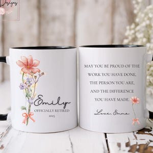 May include: White ceramic mug with a black rim and handle. The mug features a floral design with pink flowers and green leaves. The text "Emily Officially Retired 2023" is printed on the mug. The other side of the mug has the text "May you be proud of the work you have done, the person you are, and the difference you have made. Love, Emma."