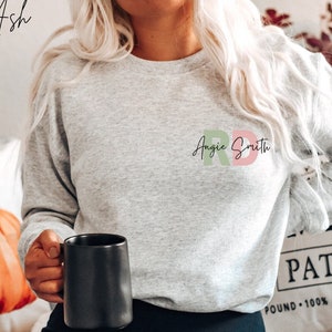May include: A gray sweatshirt with the text "Angie Smith" and the letters "RBD" in pink, green, and red. The sweatshirt is being worn by a person holding a black mug.
