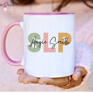 May include: White ceramic mug with a pink rim and handle. The mug has the text "Angie Smith SLP" in colorful letters.