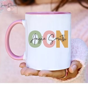 May include: White ceramic mug with a pink rim and handle. The mug has a design of the letters "OCN" in green, pink, and yellow with the text "Angie Smith" in black cursive writing.
