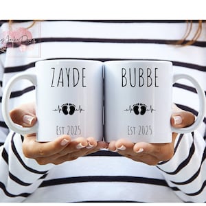 May include: Two white ceramic mugs with black handles. The mug on the left says "ZAYDE EST 2025" with a heartbeat line and two footprints. The mug on the right says "BUBBE EST 2025" with a heartbeat line and two footprints.
