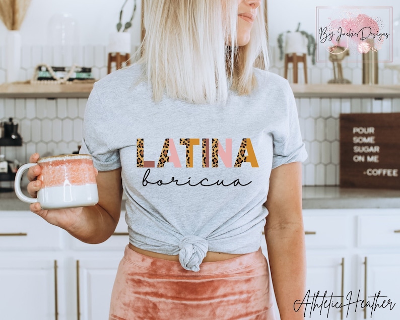K&ouml;nnte beinhalten: Graues T-Shirt mit dem Text "LATINA boricua" in bunten Buchstaben mit Leopardenmuster. Das Shirt ist in der Taille gebunden und das Model tr&auml;gt einen rosa Rock.