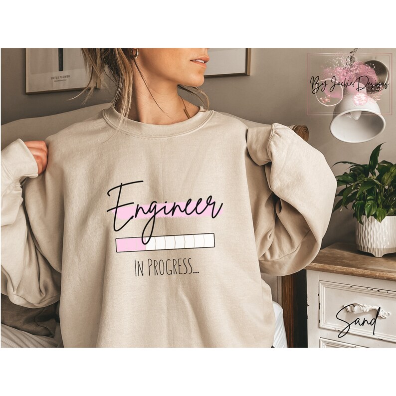 May include: Sand-colored sweatshirt with the text "Engineer" in a black script font, with a pink loading bar graphic, and the words "IN PROGRESS..." below. The sweatshirt has long sleeves.