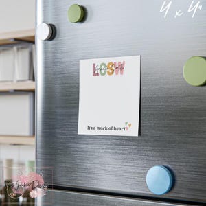 May include: A white 4x4 inch magnet with the text "LOOW Amanda Smith" in pastel colors. Below the text is the phrase "It's a work of heart" with small heart graphics. The magnet is attached to a stainless steel refrigerator.