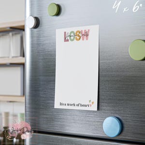 May include: A white notepad with the text "LOOSW Amanda Smith" in colorful letters is attached to a refrigerator with magnets. The notepad has the phrase "It's a work of heart" at the bottom, with small heart graphics. The notepad measures 4 x 6 inches.