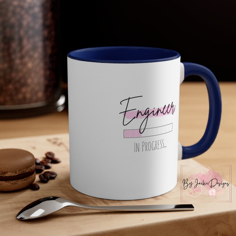 May include: White ceramic coffee mug with a blue rim and handle. The mug has a pink and black graphic that reads "Engineer in progress..."