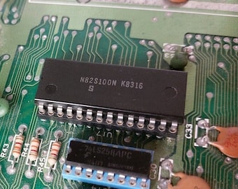 Può includere: Primo piano di una scheda a circuito verde con diversi circuiti integrati, tra cui un chip nero etichettato "N82S100N K8316 S" e un chip blu etichettato "74LSZ5HAPC".
