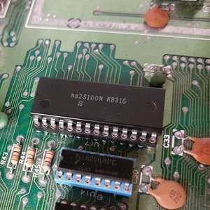 Op de afbeelding: Close-up van een groene printplaat met verschillende geïntegreerde schakelingen, waaronder een zwarte chip met het label "N82S100N K8316 S" en een blauwe chip met het label "74LSZ5HAPC".