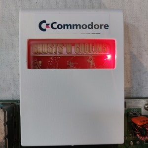 Può includere: Una cartuccia di gioco Commodore bianca con uno schermo rosso che mostra il testo "Ghosts 'n Goblins".