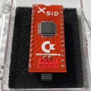 Puede incluir: Una placa de circuito impreso rectangular roja etiquetada como X-SID y commodore con un chip de circuito integrado negro central. La placa presenta un logotipo blanco, una pegatina roja que dice AUDIO BOOST y dos filas de pines metálicos en una caja.