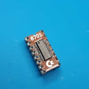 May include: A small, rectangular brown circuit board with the text "8701 TAURUS" and the Commodore logo. The board has a black rectangular chip in the center and silver pins around the edges. The background is a solid blue.