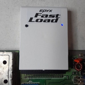 May include: A white Epyx Fast Load cartridge with a black button and a blue LED light. The cartridge is plugged into a green circuit board.