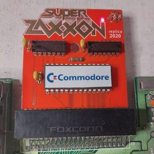 Puede incluir: Una placa de circuito roja con el texto "SUPER ZAXON" en letras doradas y el logotipo de Commodore. La placa tiene varios componentes electrónicos, incluyendo chips y un componente Foxconn. También es visible el texto "replica 2020".