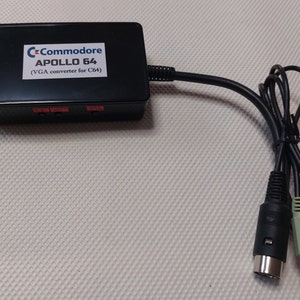 Peut inclure: Un convertisseur VGA Commodore Apollo 64 noir pour C64 avec un câble noir et deux connecteurs. Le convertisseur porte une étiquette indiquant "Commodore Apollo 64 (convertisseur VGA pour C64)".