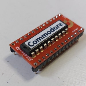 Op de afbeelding: Een rode printplaat met een zwarte chip waarop "Commodore" in witte letters staat. De printplaat heeft aan elke kant een rij gouden pinnen. Op de printplaat staat het nummer "251641-02" gedrukt.