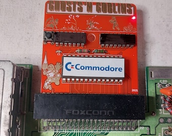 Può includere: Una scheda di circuito rosso con il testo "GHOSTS 'N GOBLINS" stampato in oro. La scheda ha un connettore nero con il testo "FOXCONN" stampato su di esso. La scheda ha anche un adesivo bianco con il testo "C-Commodore" stampato su di esso. La scheda ha una luce LED rossa nell'angolo in alto a destra. La scheda ha una data di "2021" stampata nell'angolo in basso a destra.