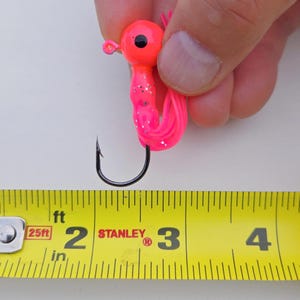 May include: A bright pink and orange fishing lure with a black hook. The lure has a round head and a curly tail. The lure is approximately 3 inches long.