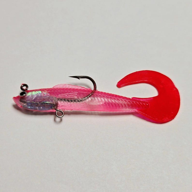 May include: A pink soft plastic fishing lure with a red tail and a black hook. The lure has a transparent body with internal detailing and a silver eye. The tail is a bright, curved shape.