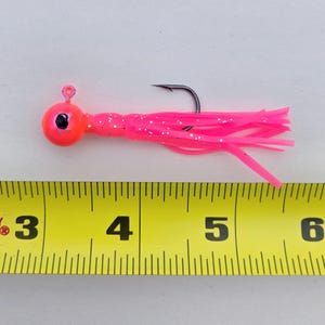 May include: A bright pink fishing lure with a round head and a long, flowing tail. The lure has a black hook and is approximately 4 inches long. The head is a vibrant orange color with a black eye. The tail is made of thin, flexible strands.