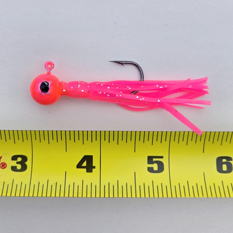 May include: A bright pink fishing lure with a round head and a long, flowing tail. The lure has a black hook and is approximately 4 inches long. The head is a vibrant orange color with a black eye. The tail is made of thin, flexible strands.