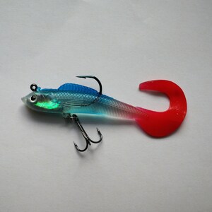 May include: A clear and blue fishing lure with a red tail and two black hooks. The lure has a fish-like shape with a curved tail and is designed to attract fish. The lure is designed for freshwater fishing.