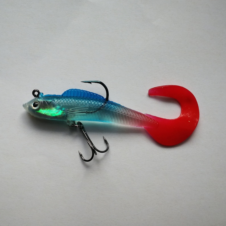 May include: A clear and blue fishing lure with a red tail and two black hooks. The lure has a fish-like shape with a curved tail and is designed to attract fish. The lure is designed for freshwater fishing.