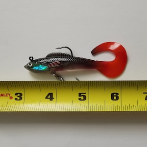 May include: A black and red fishing lure with a curved red tail and a silver hook. The lure is approximately 6 inches long and has iridescent accents on the head. The lure is designed to mimic a small fish.