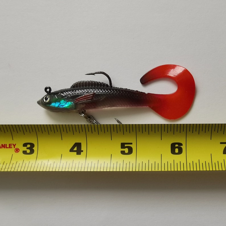 May include: A black and red fishing lure with a curved red tail and a silver hook. The lure is approximately 6 inches long and has iridescent accents on the head. The lure is designed to mimic a small fish.