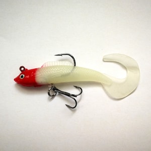 May include: A soft plastic fishing lure in the shape of a fish. The lure has a red head, a translucent body, and a curved tail. It is equipped with two treble hooks and a single hook.