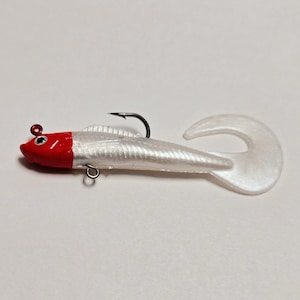 May include: A white and red fishing lure with a curved tail and a black hook. The lure has a red head and a white body with a textured surface. A small metal loop is attached to the body.