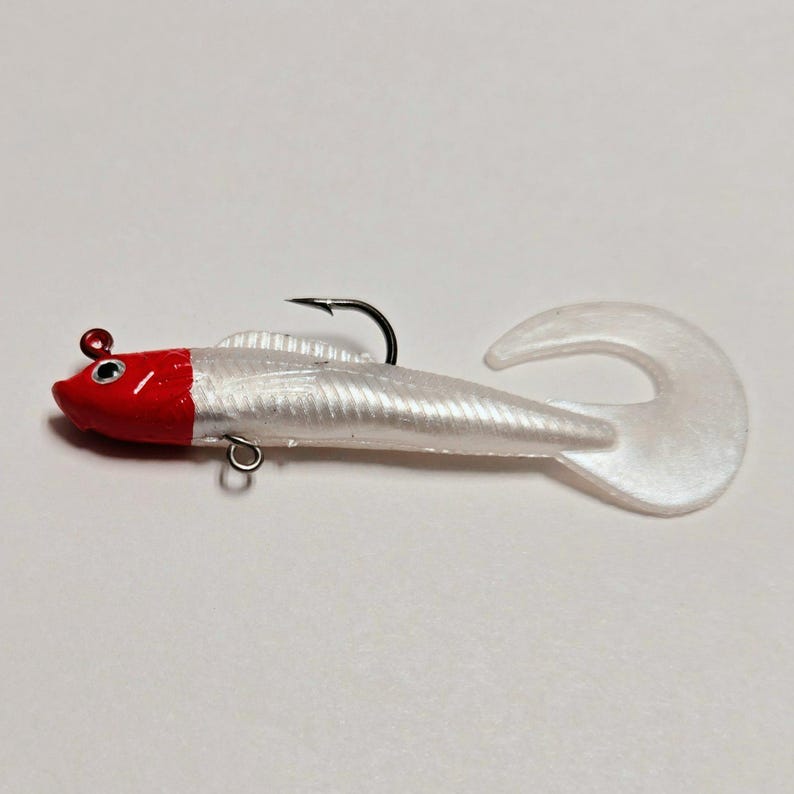 May include: A white and red fishing lure with a curved tail and a black hook. The lure has a red head and a white body with a textured surface. A small metal loop is attached to the body.