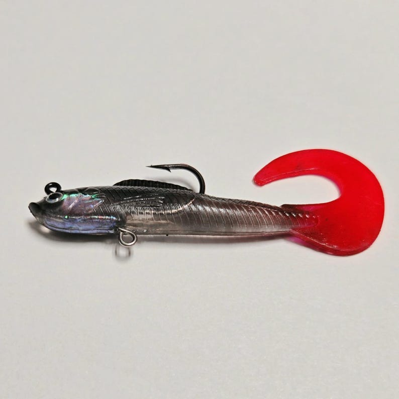 May include: A clear, fish-shaped fishing lure with a red tail. The lure has a black hook and a silver body with iridescent details. The lure is designed to attract fish.