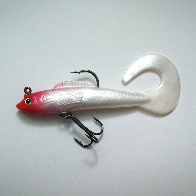 May include: A fishing lure shaped like a fish, with a red head, silver body, and a white, curved tail. It has two black hooks and a small red eye. The lure is designed to attract fish.