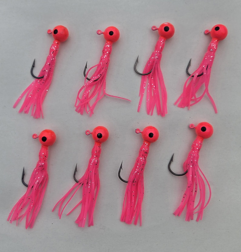 May include: Eight pink fishing jigs with orange heads and black hooks are arranged on a light background. The jigs have a teardrop-shaped head, a black hook, and a pink skirt with silver glitter.