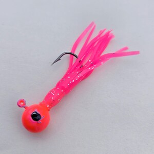 May include: A bright pink fishing lure with a round, orange head and a black hook. The lure has a cluster of pink, tentacle-like strands with silver glitter accents, designed to attract fish.
