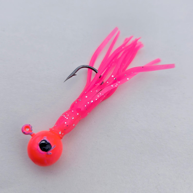 May include: A bright pink fishing lure with a round, orange head and a black hook. The lure has a cluster of pink, tentacle-like strands with silver glitter accents, designed to attract fish.