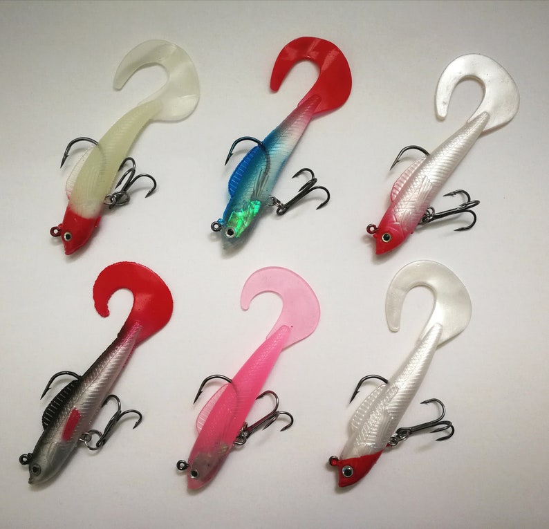 May include: Six colorful fishing lures with curved tails and treble hooks. The lures feature a variety of colors, including red, white, pink, and blue, with silver and black accents. The lures are designed to attract fish.