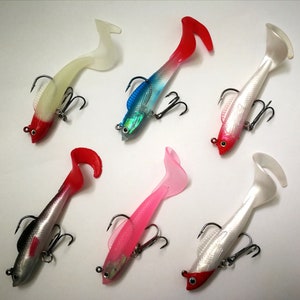 May include: Six soft plastic fishing lures in various colors, including red, white, pink, and blue. Each lure features a fish-like design with a tail and a treble hook for catching fish. The lures are designed for freshwater or saltwater fishing.