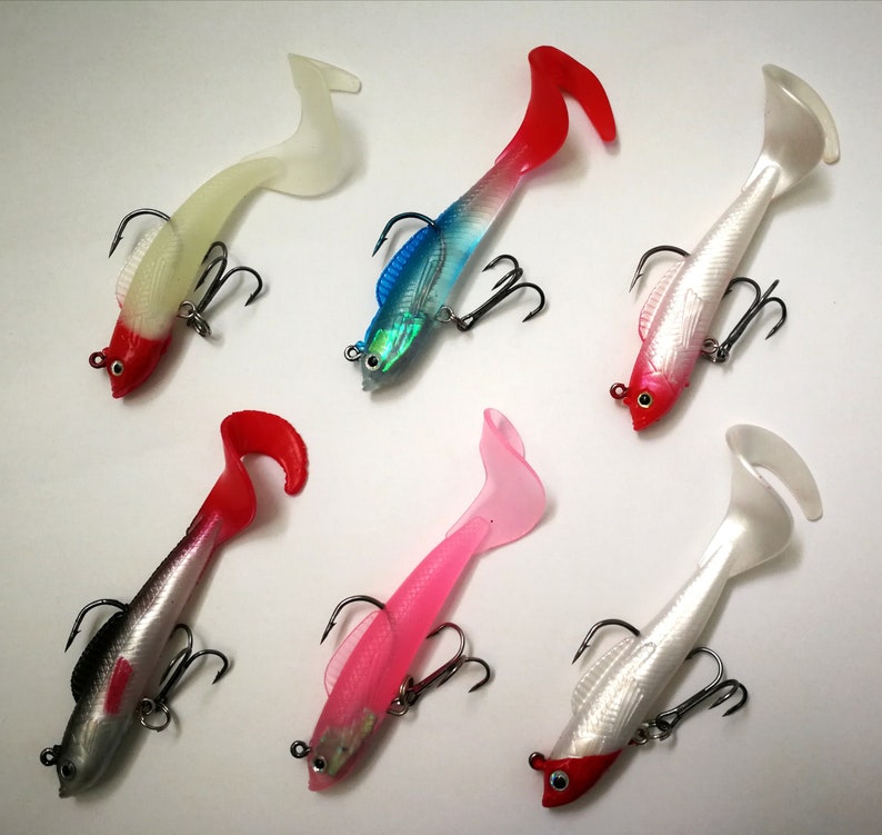 May include: Six soft plastic fishing lures in various colors, including red, white, pink, and blue. Each lure features a fish-like design with a tail and a treble hook for catching fish. The lures are designed for freshwater or saltwater fishing.
