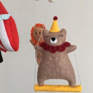 May include: A brown felt bear wearing a red and yellow circus costume is swinging on a yellow bar. The bear has a red nose and closed eyes. The bear is part of a felt mobile.