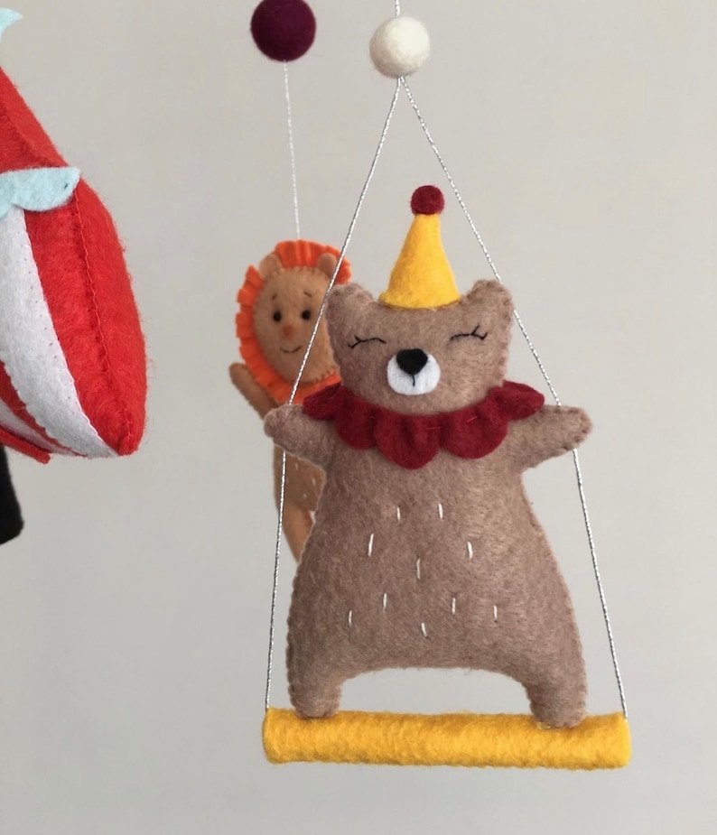 May include: A brown felt bear wearing a red and yellow circus costume is swinging on a yellow bar. The bear has a red nose and closed eyes. The bear is part of a felt mobile.