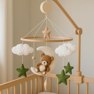 May include: A wooden baby mobile featuring a brown teddy bear sitting on a cream-colored crescent moon. The mobile includes white clouds, green stars, and a wooden star, all suspended from a wooden ring. The mobile is attached to a wooden crib.