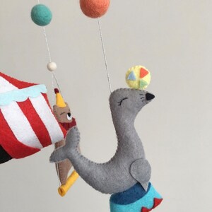 May include: A gray felt seal balancing a colorful ball on its nose. The seal is standing on a blue and red circus ring. The seal is part of a felt mobile with a red and white striped tent, a brown bear, and colorful balls.