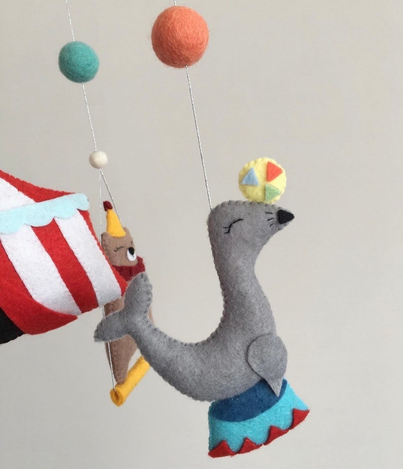 May include: A gray felt seal balancing a colorful ball on its nose. The seal is standing on a blue and red circus ring. The seal is part of a felt mobile with a red and white striped tent, a brown bear, and colorful balls.