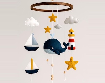 May include: A baby mobile with a nautical theme, featuring a dark blue whale, sailboats, a lighthouse, yellow stars, and white clouds. The mobile is suspended from a wooden ring and includes wooden beads. The colour palette includes navy, white, yellow, and red.